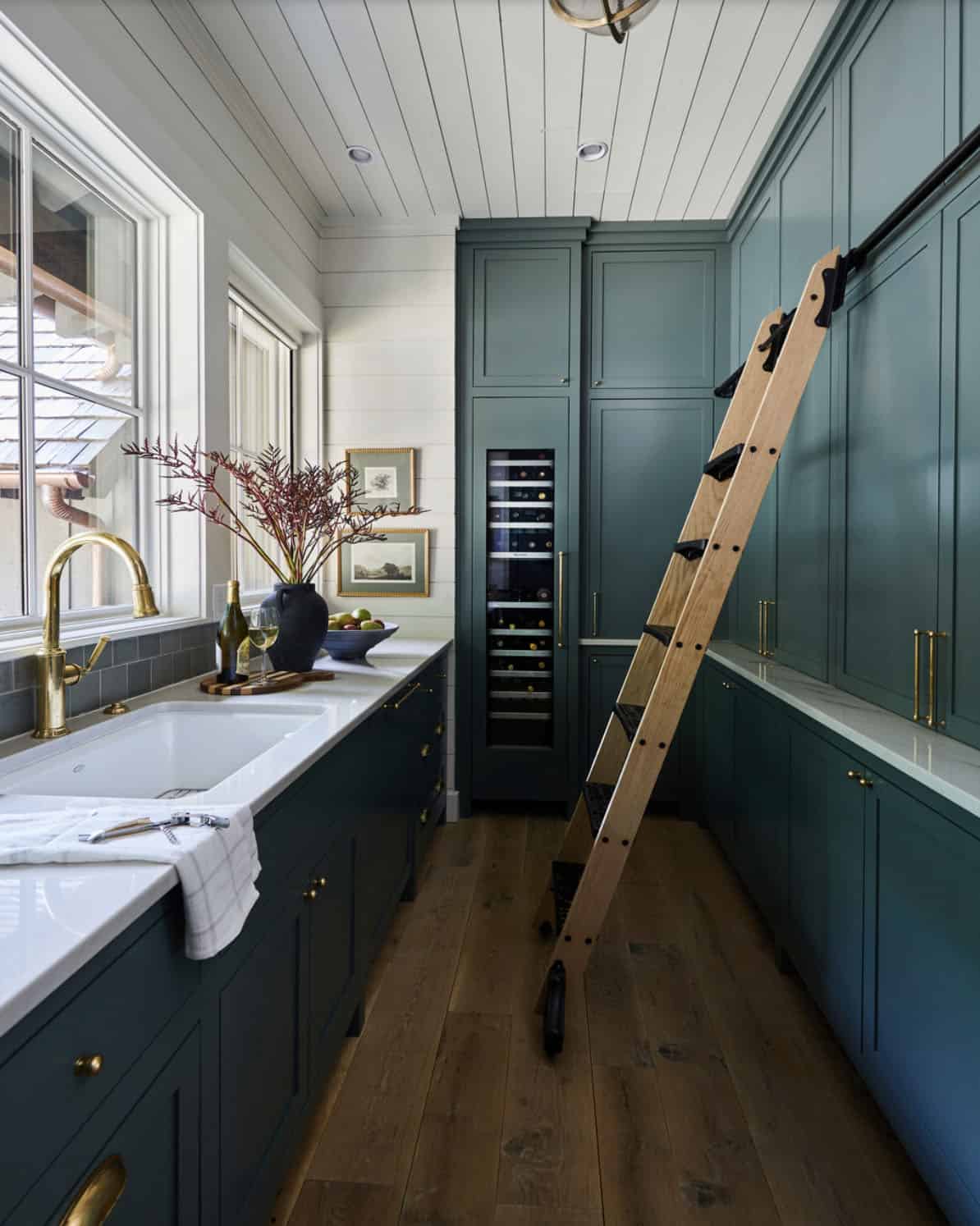 Deep teal butler's pantry with floor-to-ceiling shaker cabinetry, brass hardware, farmhouse sink, brass gooseneck faucet, wine refrigerator, rolling library ladder, white shiplap ceiling, and wide plank oak floors
