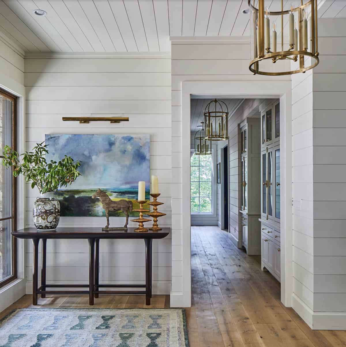 Foyer with white shiplap walls, dark wood console table styled with abstract landscape painting, bronze horse sculpture, brass candlesticks, large ceramic vase, brass picture light, vintage rug, and brass lantern pendants down hallway