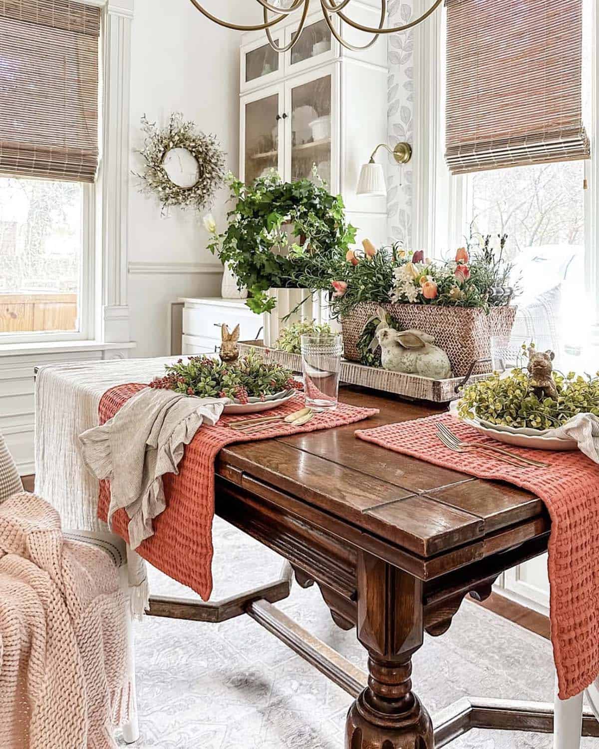 Spring breakfast nook decorating ideas with coral placemats, a linen table runner, a wicker basket centerpiece of tulips and trailing ivy, ceramic bunnies, and a dried wreath on botanical wallpaper