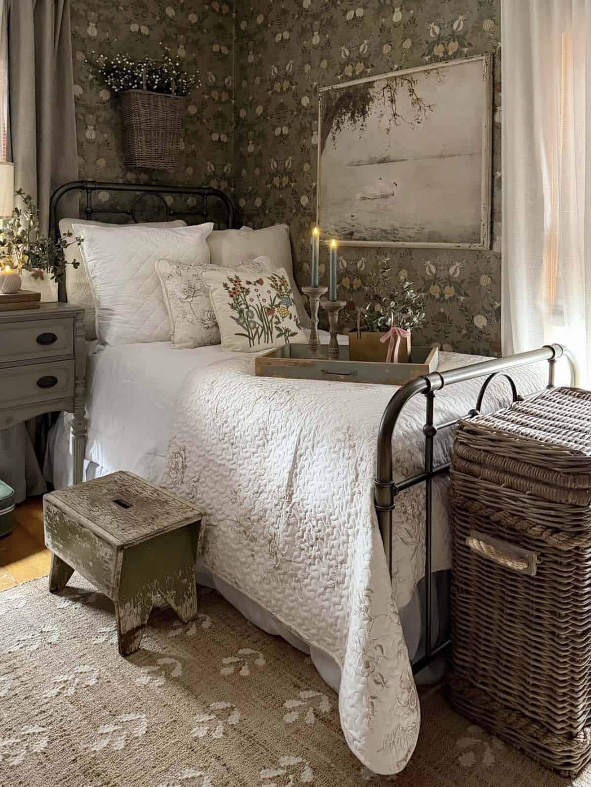 Cozy spring bedroom with sage green botanical wallpaper, black iron bed, white quilted bedding, embroidered floral pillows, vintage swan painting, taper candles, gray painted nightstand and wicker storage basket