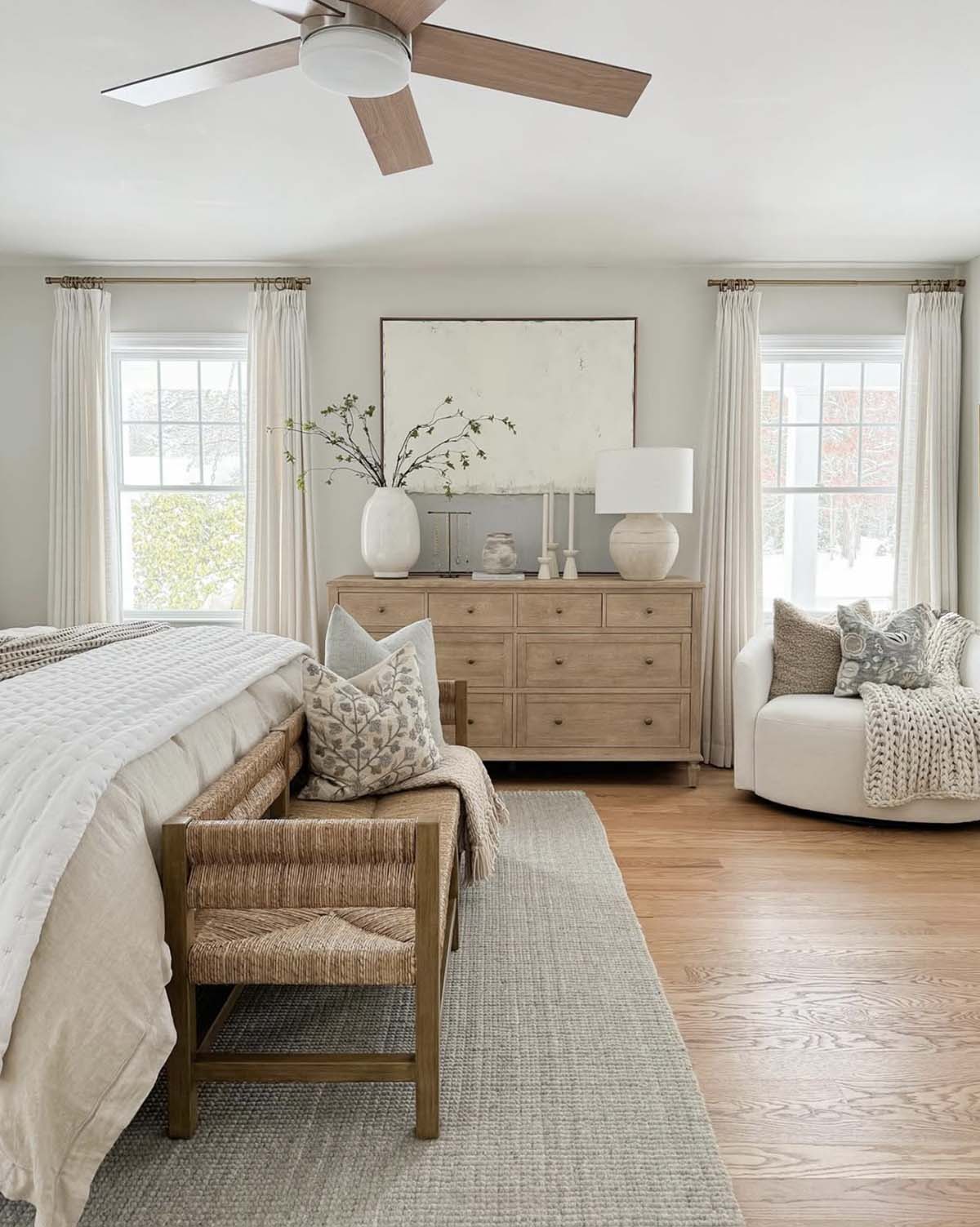 Neutral spring bedroom with white quilted bedding, woven rush bench, oak dresser, white ceramic vases with branches, cream swivel chair and linen drapery panels