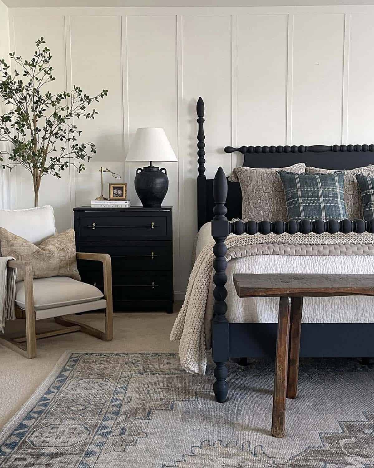 Spring bedroom with black spindle four-poster bed, white board and batten wall, neutral quilted bedding, teal plaid accent pillow, black nightstand with terracotta lamp, rustic wood bench, linen accent chair, vintage blue rug and tall olive branch stems