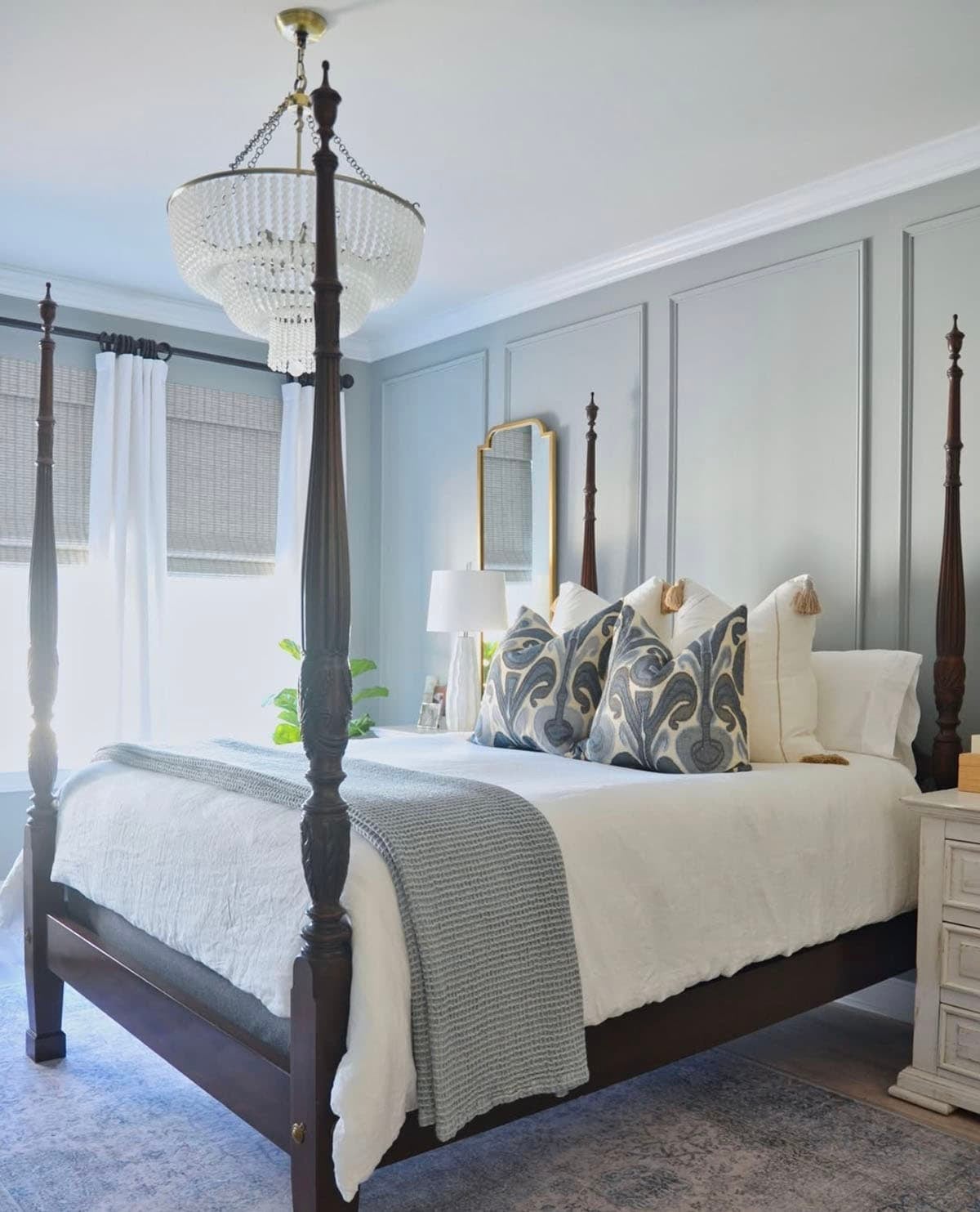 Spring bedroom with dark wood four-poster bed, soft gray paneled accent wall, white linen bedding, blue paisley accent pillows, gray waffle knit throw, crystal beaded chandelier, gold framed mirror, woven roman shades and vintage blue area rug