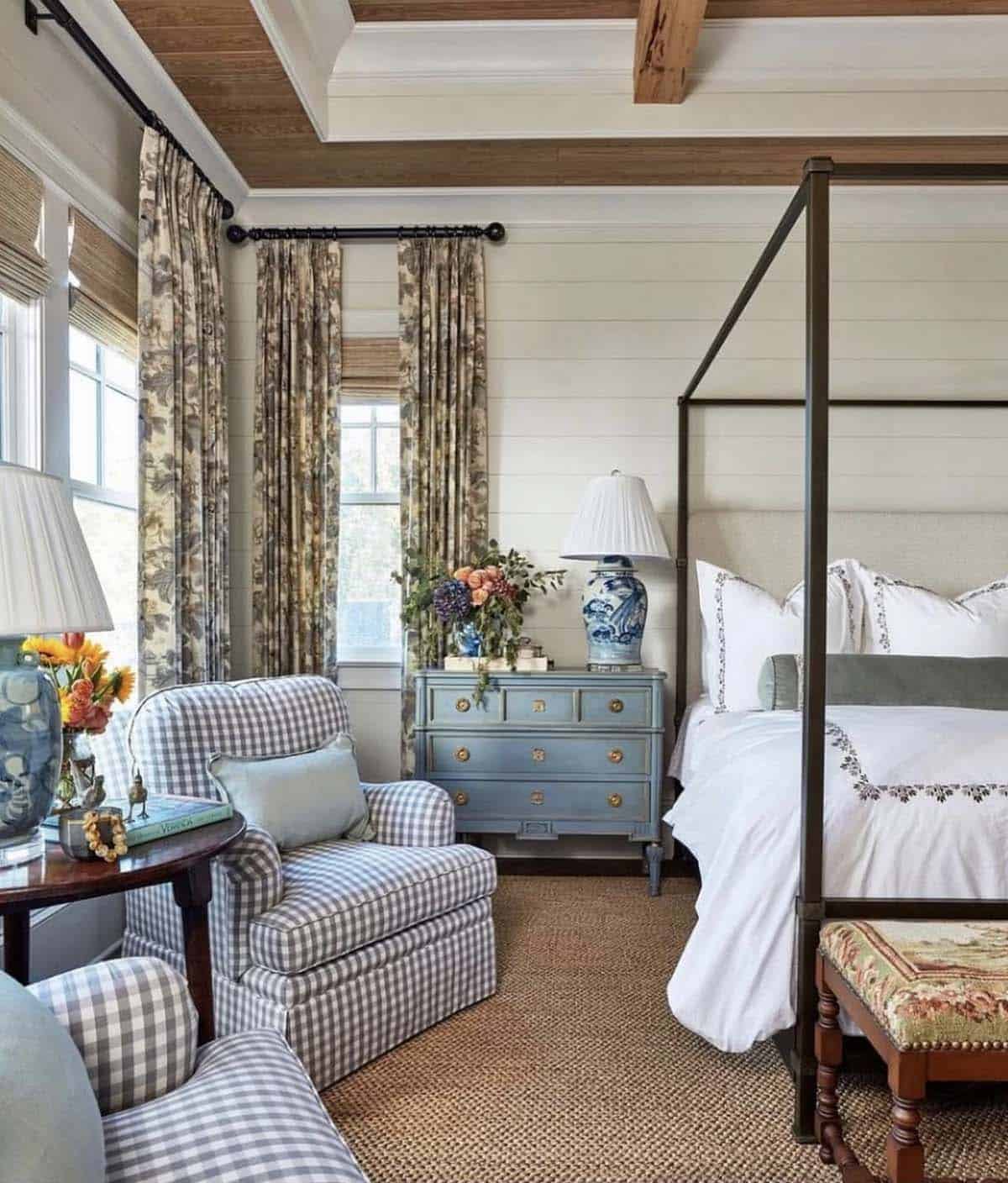 Spring bedroom with bronze four-poster bed, white embroidered bedding, blue painted chest nightstand, chinoiserie ginger jar lamp, floral toile curtains, blue gingham armchairs, wood coffered ceiling, shiplap walls and sisal area rug