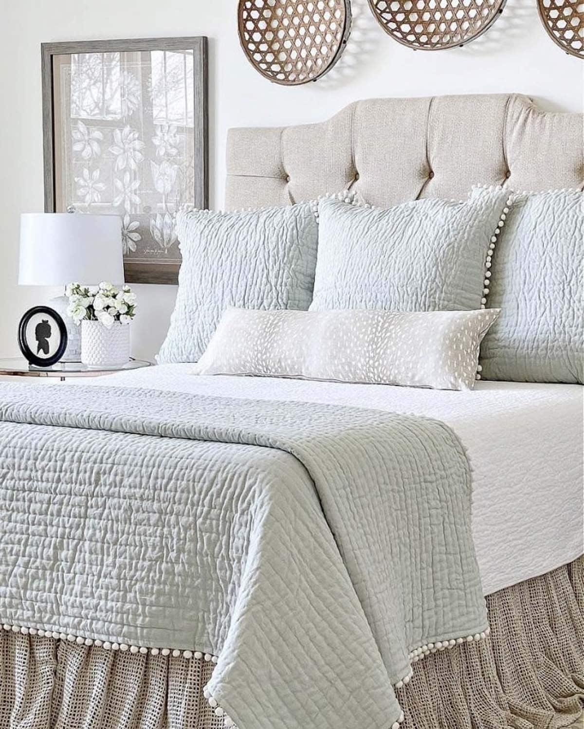 Soft spring bedroom with linen tufted headboard, powder blue quilted bedding with pom pom trim, white fawn print lumbar pillow, ruffled burlap bed skirt, round woven cane wall decor and white drum table lamp