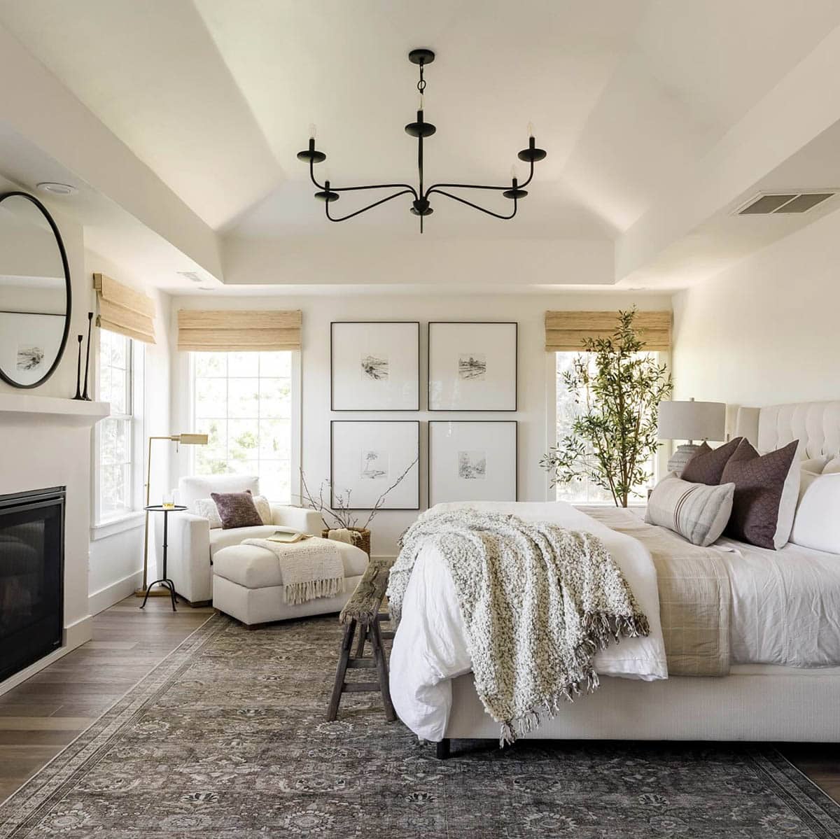 Bright transitional bedroom with vaulted tray ceiling, black iron chandelier, cream tufted upholstered bed, white linen bedding with chunky knit throw, plum velvet pillows, four-panel sketch art grid above headboard, woven grass roman shades, vintage Persian rug, white chaise lounge, fireplace, and potted olive tree