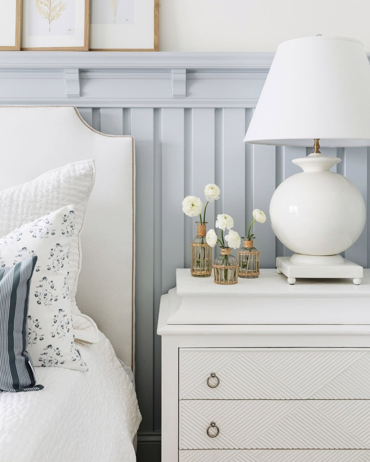 Spring bedroom detail with white upholstered wingback headboard, floral and ticking stripe pillows, white quilted bedding, gray painted board and batten wainscoting, white geometric nightstand with ring pulls, round white ceramic table lamp, and rattan-wrapped bud vases with white ranunculus
