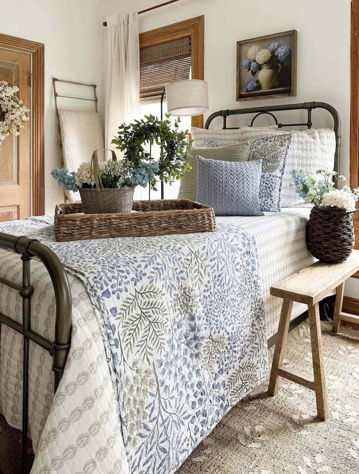 Spring farmhouse bedroom with olive green iron bed, blue and white botanical leaf quilt, layered blue floral pillows, wicker tray with greenery wreath and blue flowers, rustic wood bench, and framed hydrangea painting