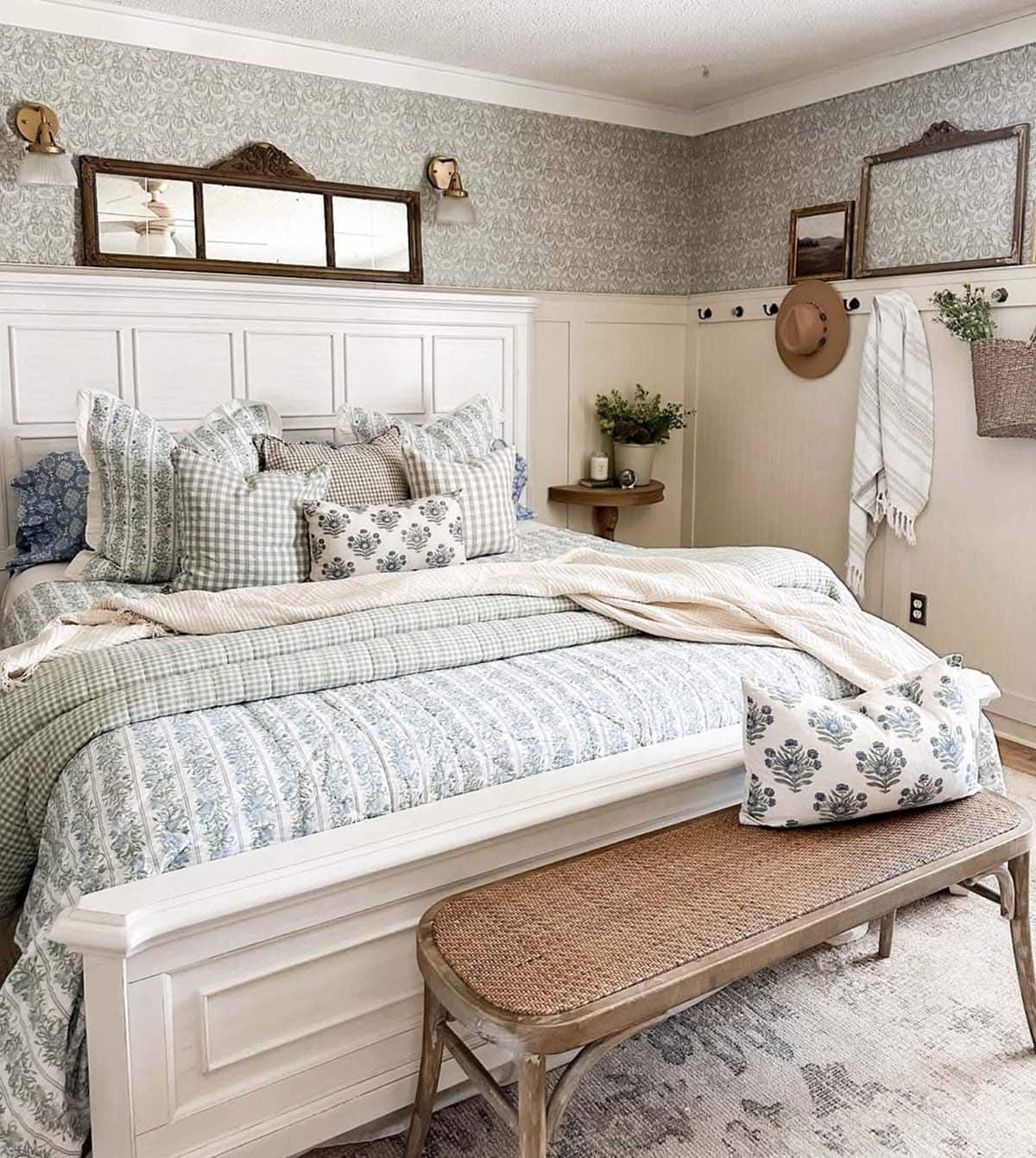 Cottage bedroom with sage green wallpaper, white panel bed, mixed blue floral and gingham bedding, and cane bench