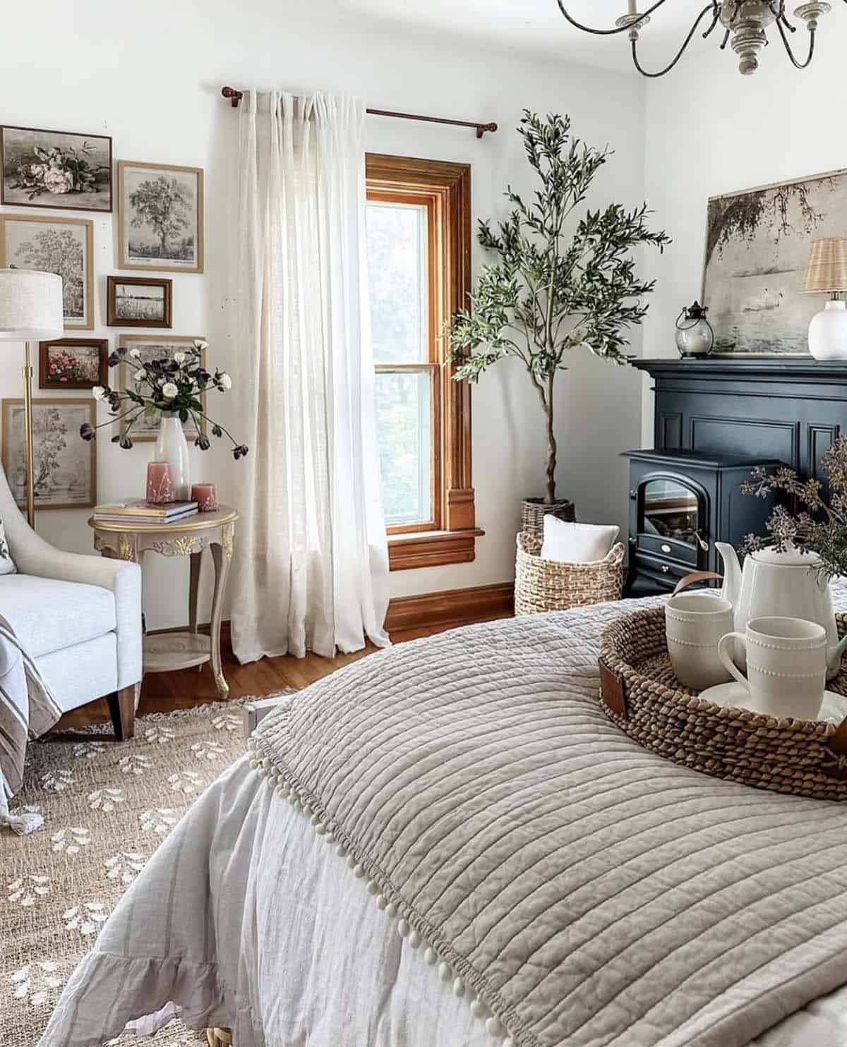 cottage-style bedroom with linen quilted bedding and pom-pom trim, wicker breakfast tray, navy painted fireplace, olive tree in woven basket, French gilt side table, botanical gallery wall in mixed gilt frames, sheer white curtains on wood curtain rod, and jute rug
