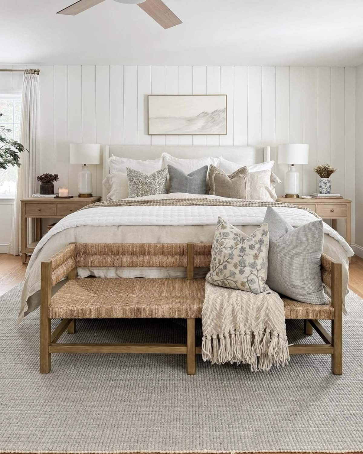 Neutral spring bedroom with linen upholstered headboard, white quilted bedding, layered floral and gray accent pillows, woven rush bench, oak nightstands, shiplap accent wall and landscape artwork