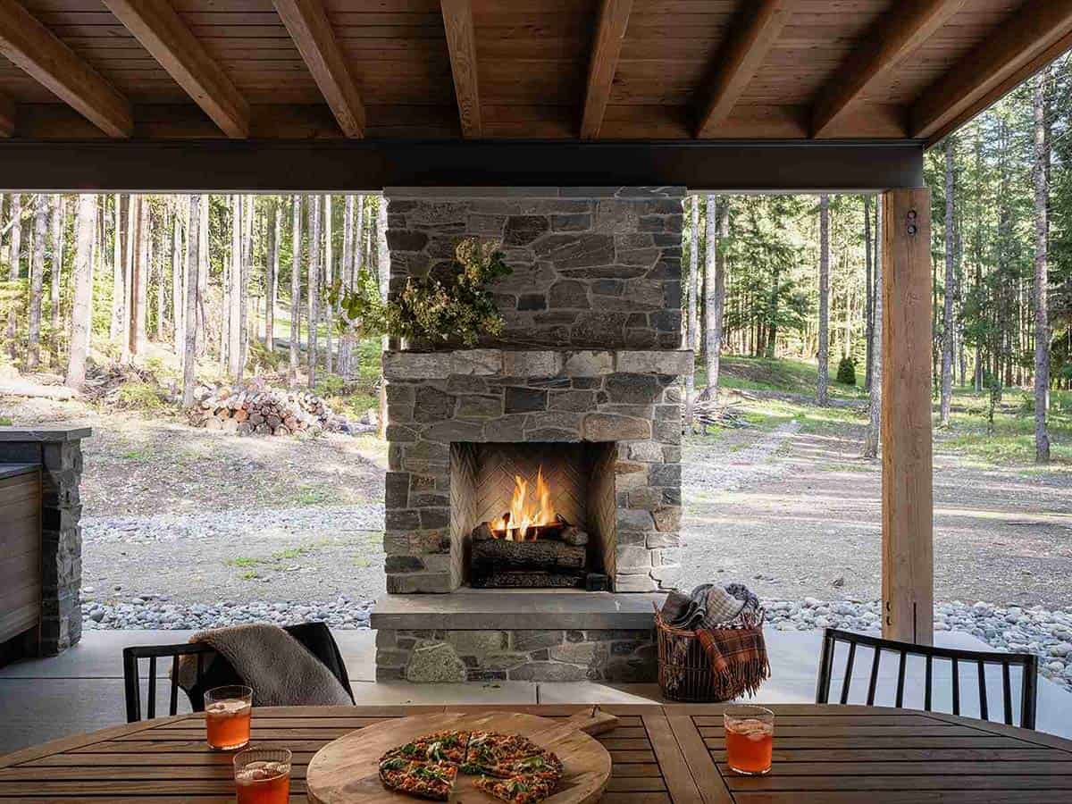 Covered outdoor patio with stone fireplace, wood beam ceiling, and dining table surrounded by pine forest, Greenwater, Washington