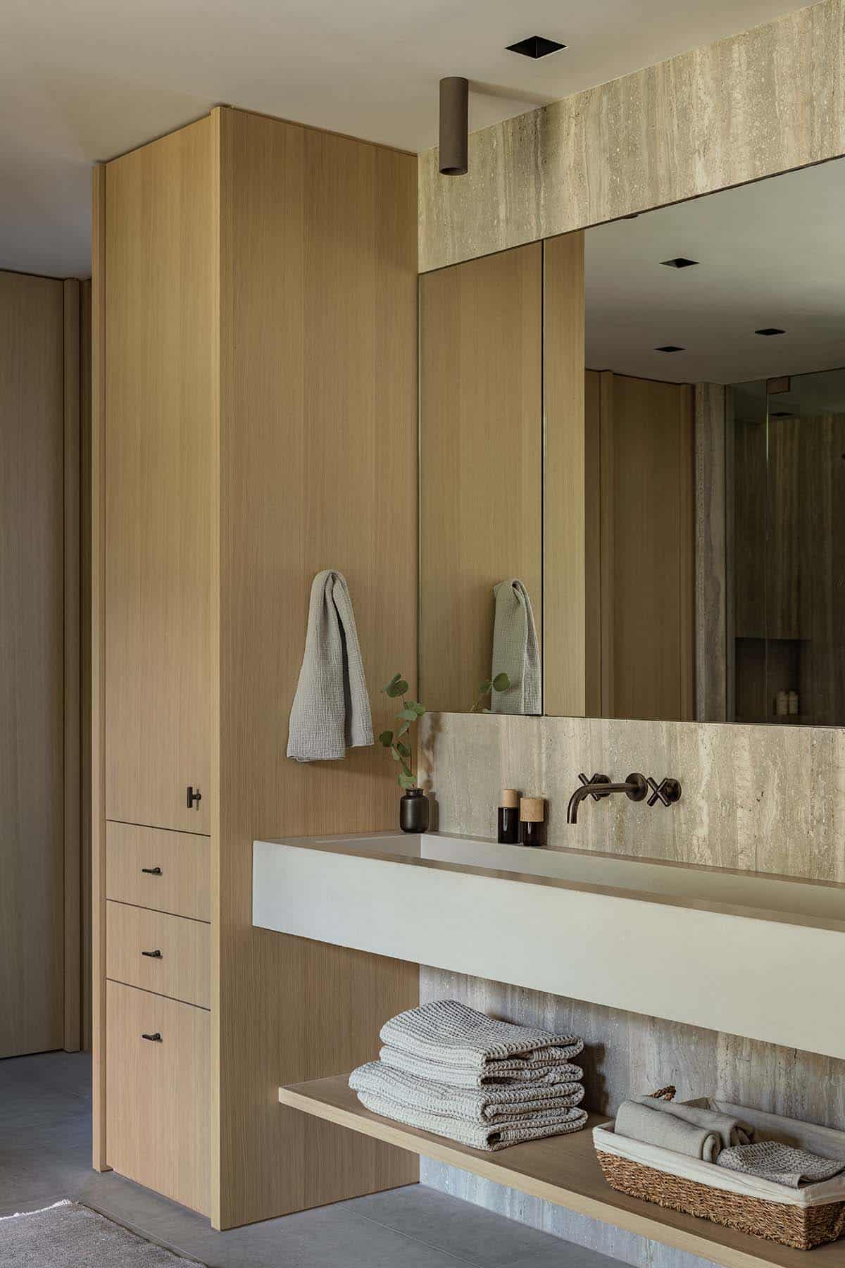 Minimalist bathroom vanity with oak cabinetry, travertine backsplash, and wall-mount faucet