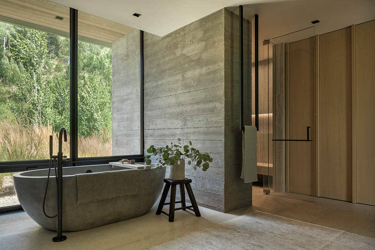Freestanding concrete soaking tub beside floor-to-ceiling windows overlooking aspen grove