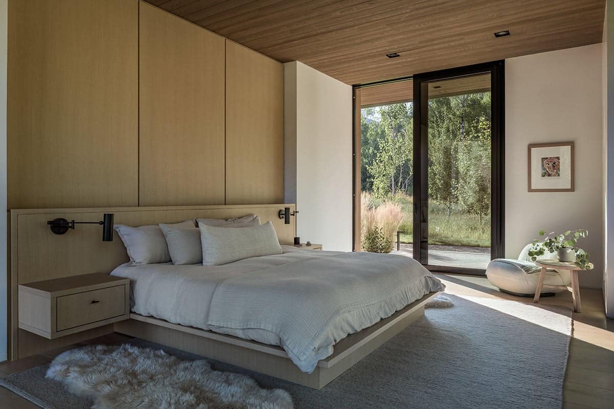 Guest bedroom with built-in oak headboard wall, black swing-arm sconces, and patio door to meadow