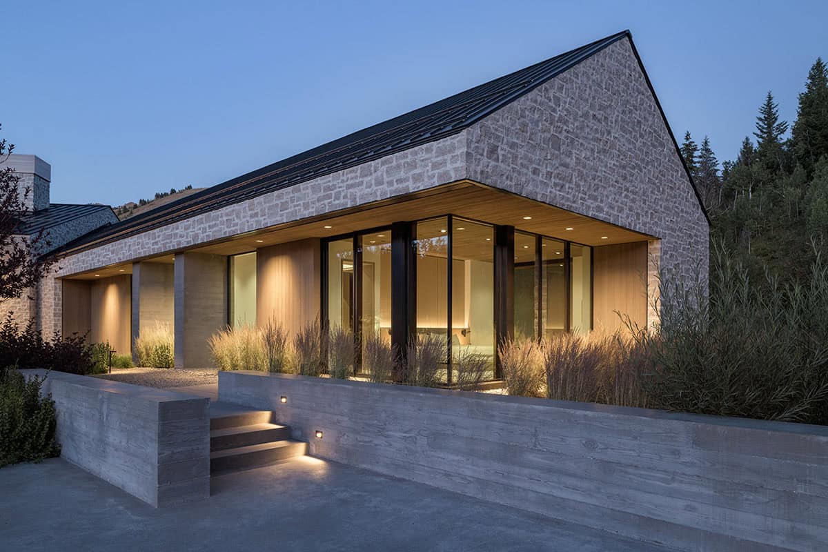 Stone gable estate at dusk with illuminated floor-to-ceiling windows, wood soffit, and concrete steps