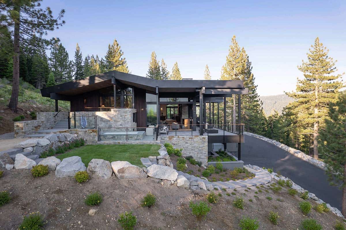Rear exterior of ski retreat at dusk with stone cladding, spa, deck, and pine forest