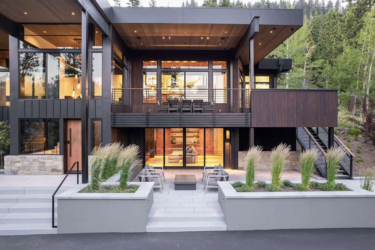 Rear exterior at dusk with lit patio, fire pit seating, upper dining deck, and glass walls