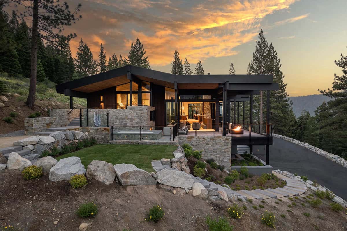 Modern mountain home at sunset with stone spa, fire pit deck, and glowing interior