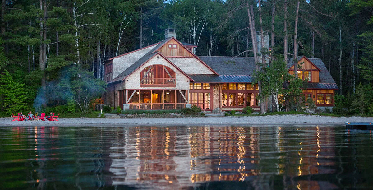 Classic New England lake house exterior nestled among mature trees on the private sandy beachfront of Lake Sunapee, Newbury, New Hampshire &mdash; shingle siding and stone details visible through the canopy