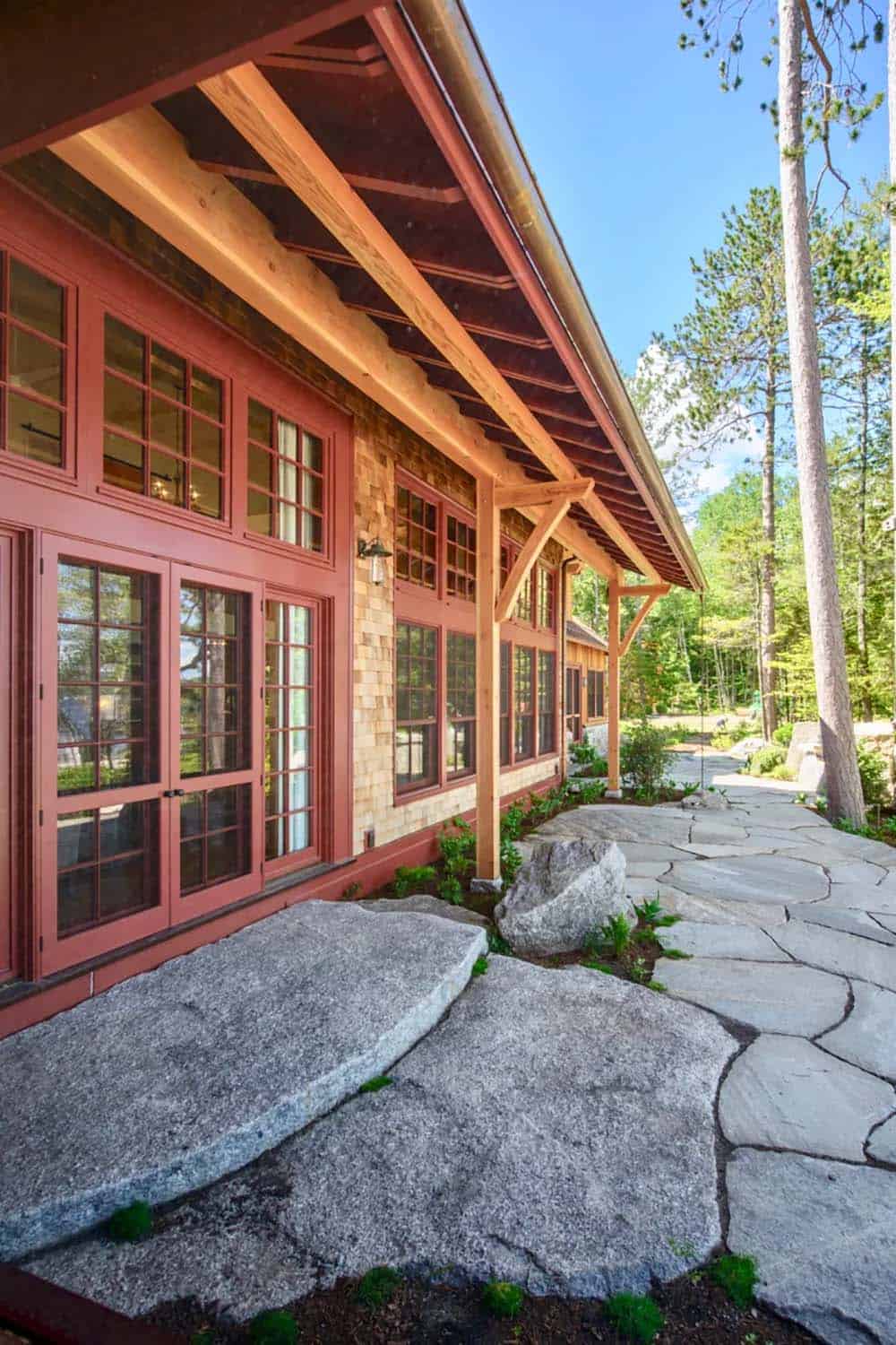 Exterior detail of the Lake Sunapee lake house showing cedar shingle siding, red-trimmed French doors, a timber frame roof overhang, and a natural stone pathway leading through native plantings