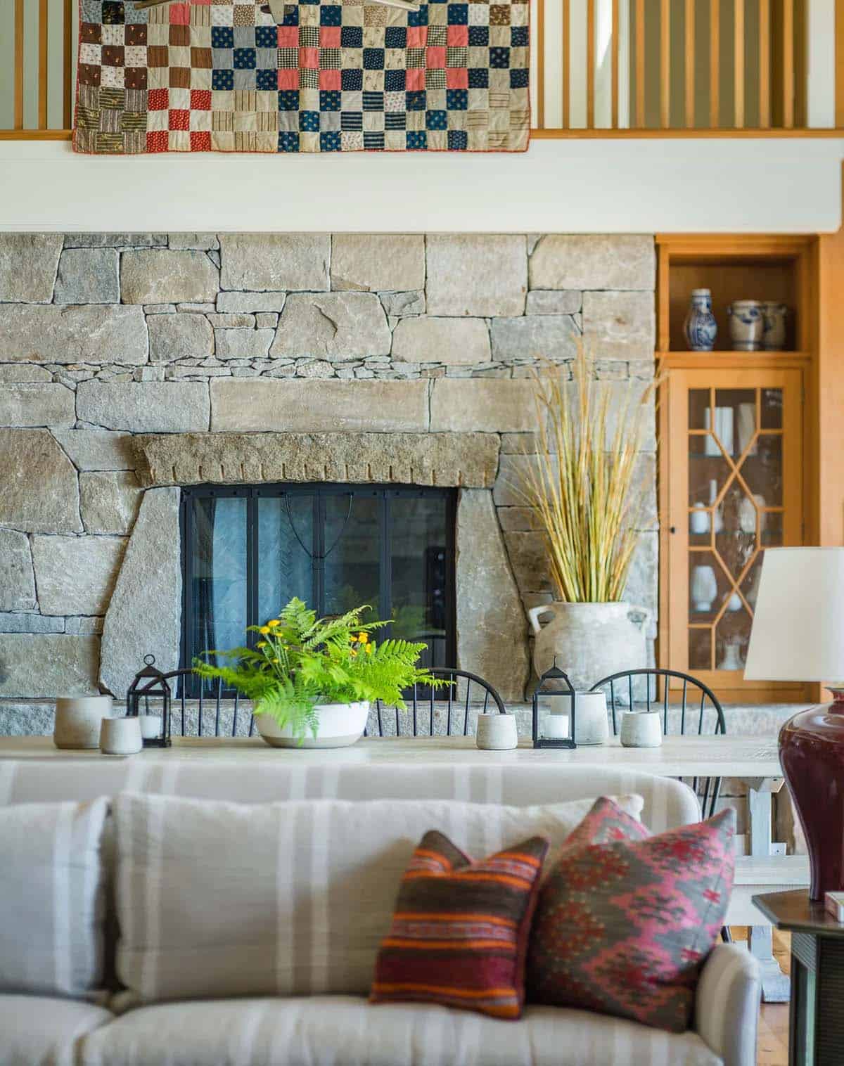 Double-sided stone fireplace at the end of the great room &mdash; local fieldstone surround, thick wood mantel, and a sinuous second-floor railing visible above, Lake Sunapee New Hampshire lake house