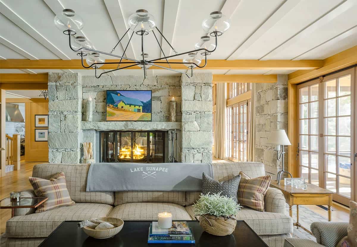 The pub area of the Lake Sunapee New Hampshire lake house &mdash; a stone-clad bar wall, built-in shelving with colorful curated bar accessories, cozy seating, and a fireplace at one end of the great room
