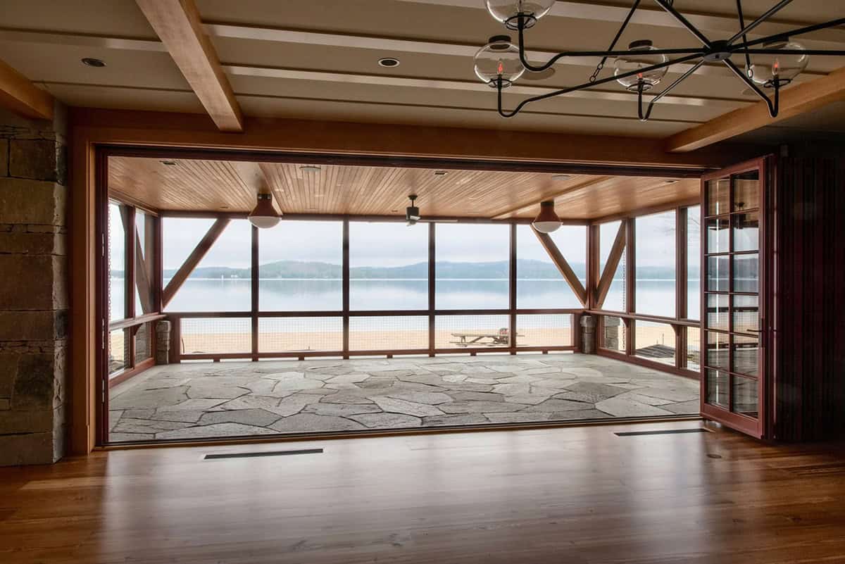 Wide view of the screened porch showing the full sitting arrangement, painted wood columns, and the lake visible beyond the screen &mdash; a classic New England summer living room