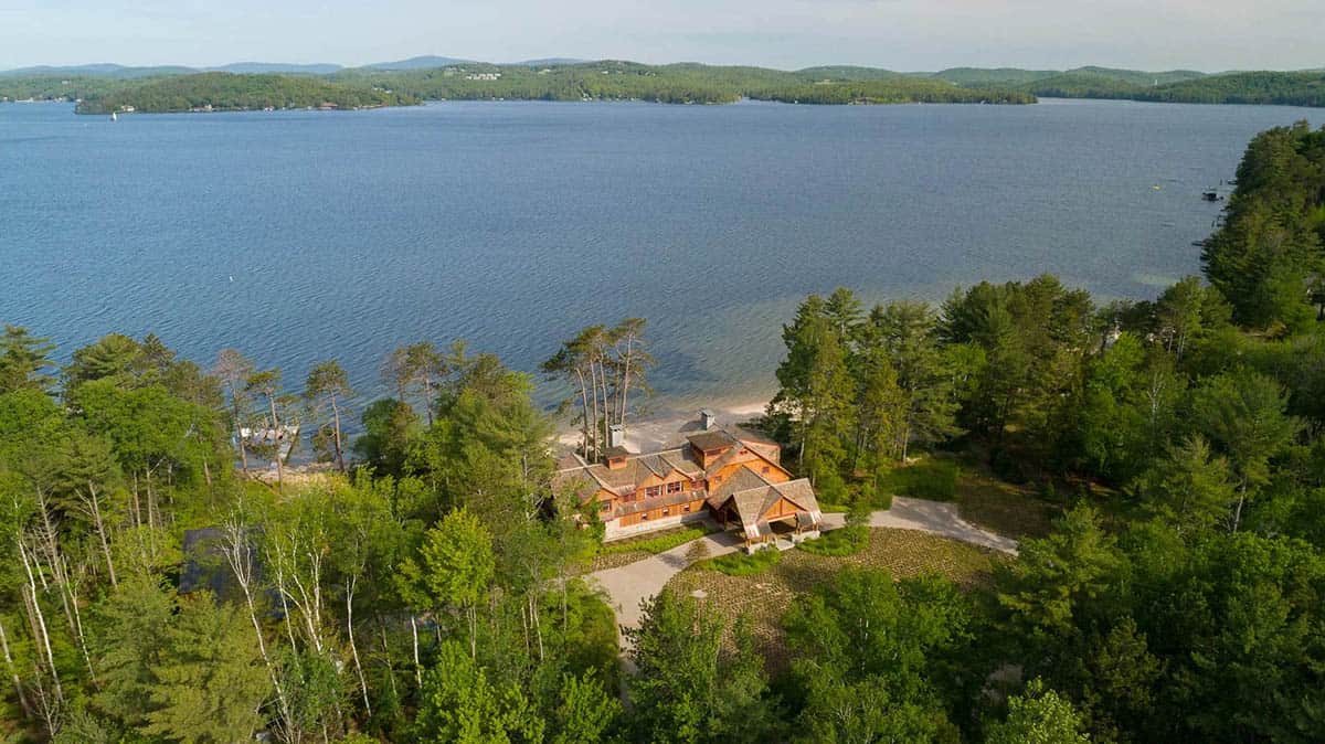 Aerial view of the classic New England lake house compound on Lake Sunapee, New Hampshire &mdash; the main house, barn, and beach cabin visible from above, surrounded by native trees and a two-acre meadow with direct access to the private sandy beach