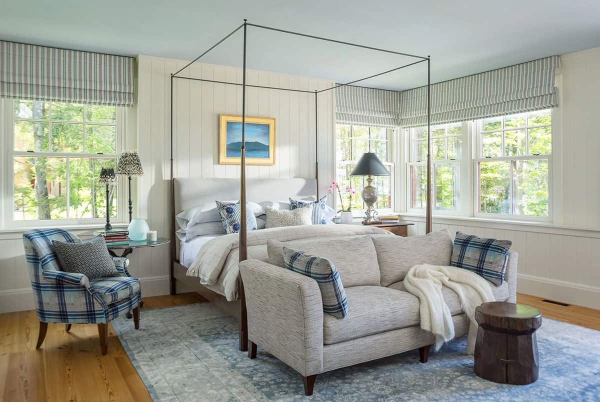 Second view of the primary bedroom in the Lake Sunapee summer house &mdash; plaid bedding, antique nightstands, a painted wood ceiling, and a window seat offering views across the private sandy beach