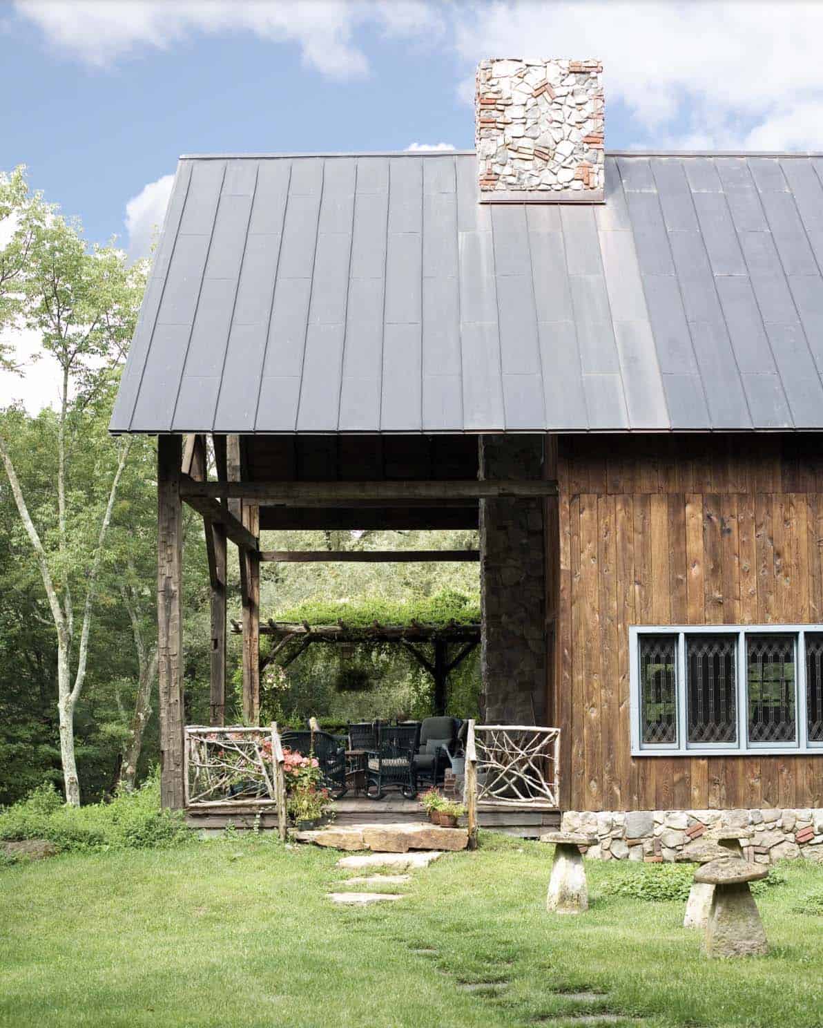 Covered porch with rustic twig railing, stone chimney, and metal roof on barn retreat