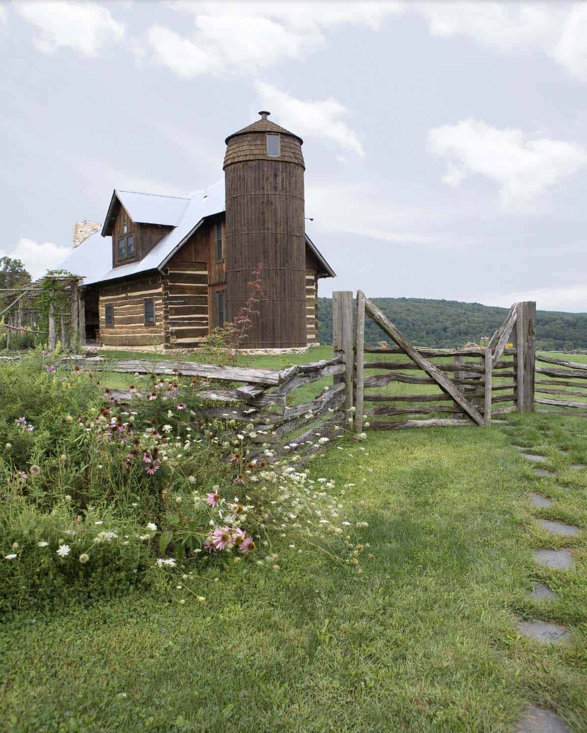 Log cabin with wooden silo, split-rail fence gate, and wildflower meadow in foreground
