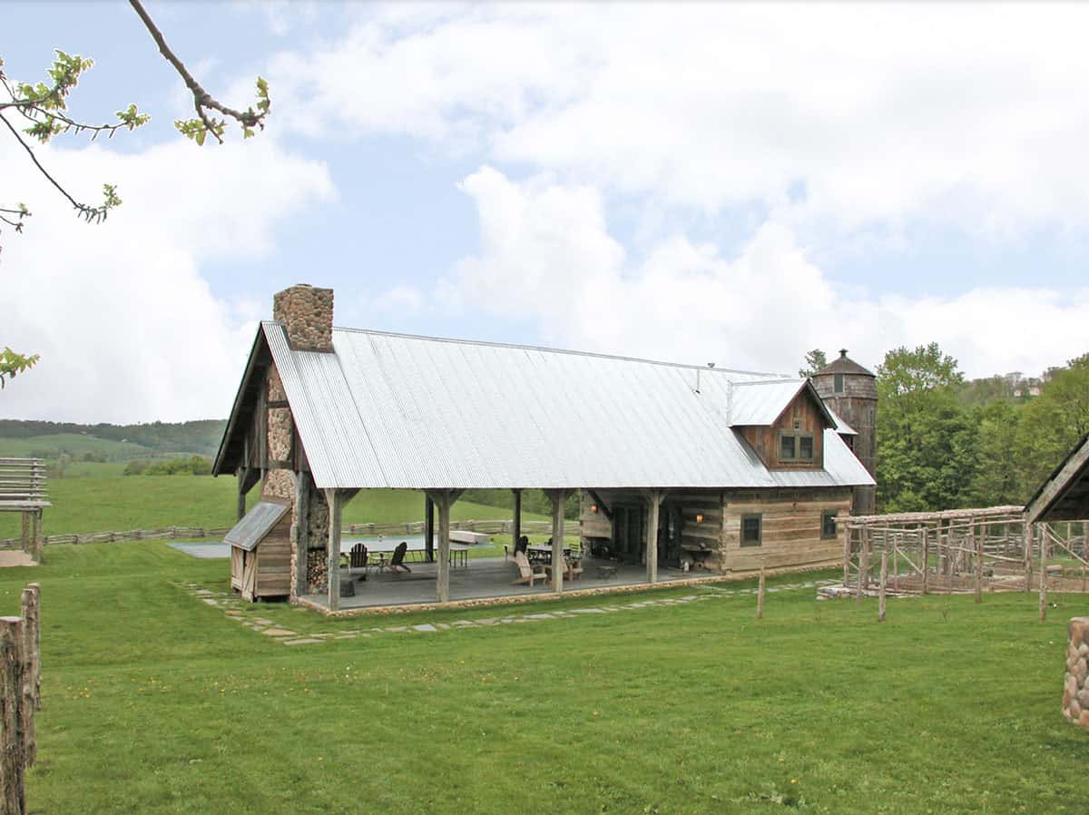 Wide view of log cabin with covered porch, stone chimney, wooden silo, and rolling green fields