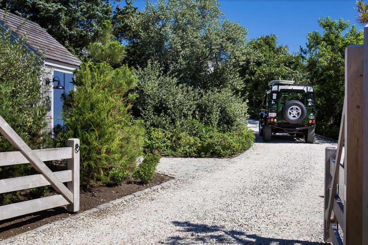 Gravel driveway with wooden gate, blue-shuttered shed, and lush coastal plantings at a Massachusetts beach house