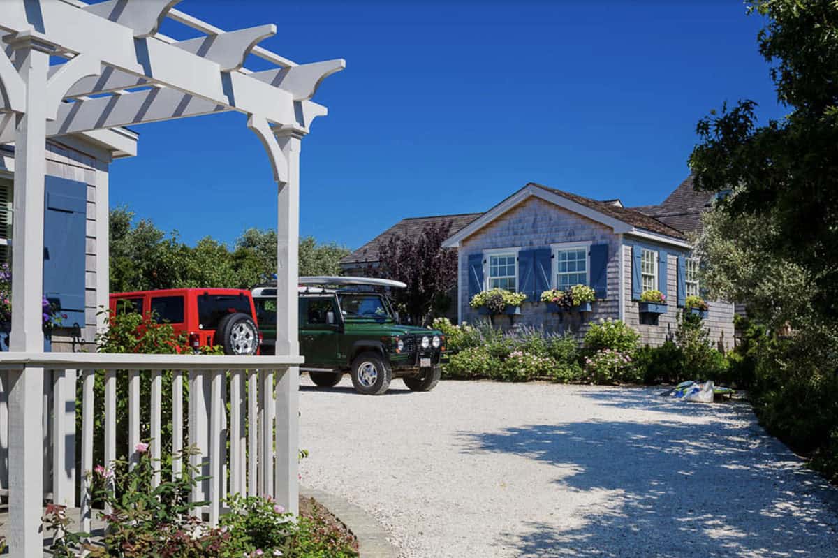 View through white pergola toward cedar shingle cottage with blue shutters and flower-filled window boxes