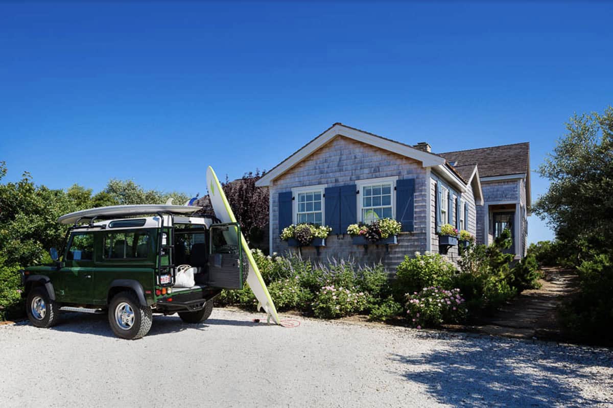 Green Land Rover with surfboard parked in front of cedar shingle cottage with blue shutters