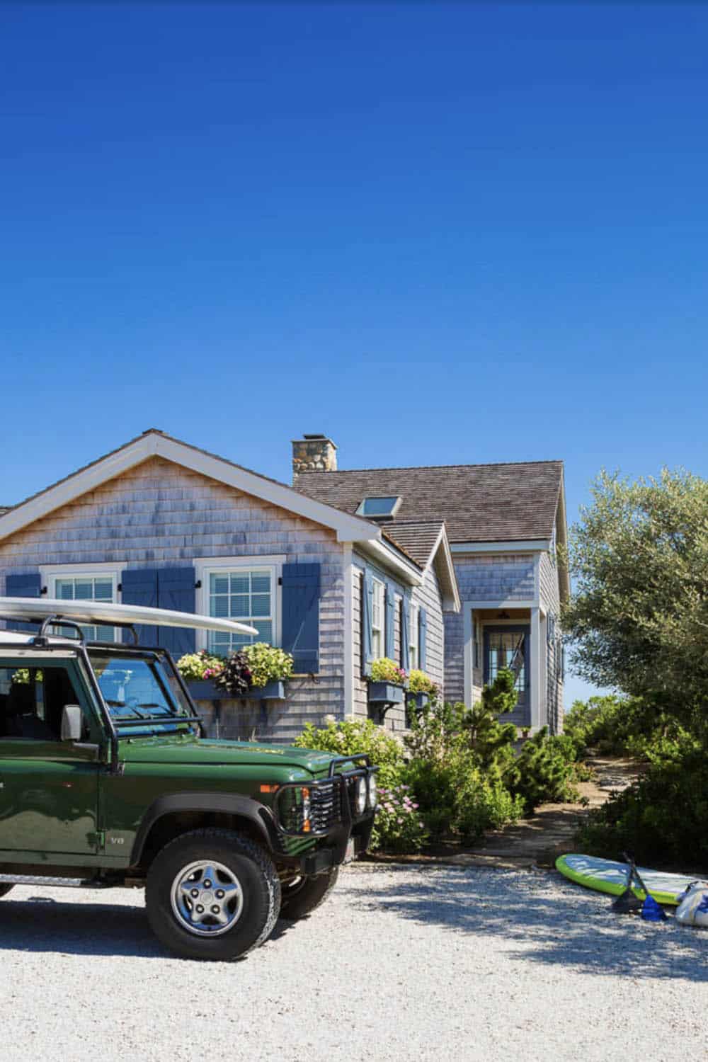 Cedar shingle beach cottage with blue shutters, stone chimney, and paddleboards on gravel drive