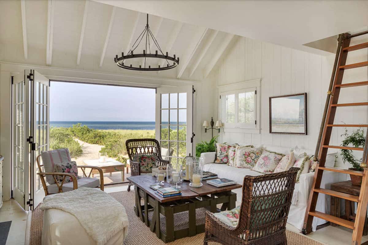 Bright living room with cathedral ceiling, wicker chairs, floral pillows, and ocean views through French doors