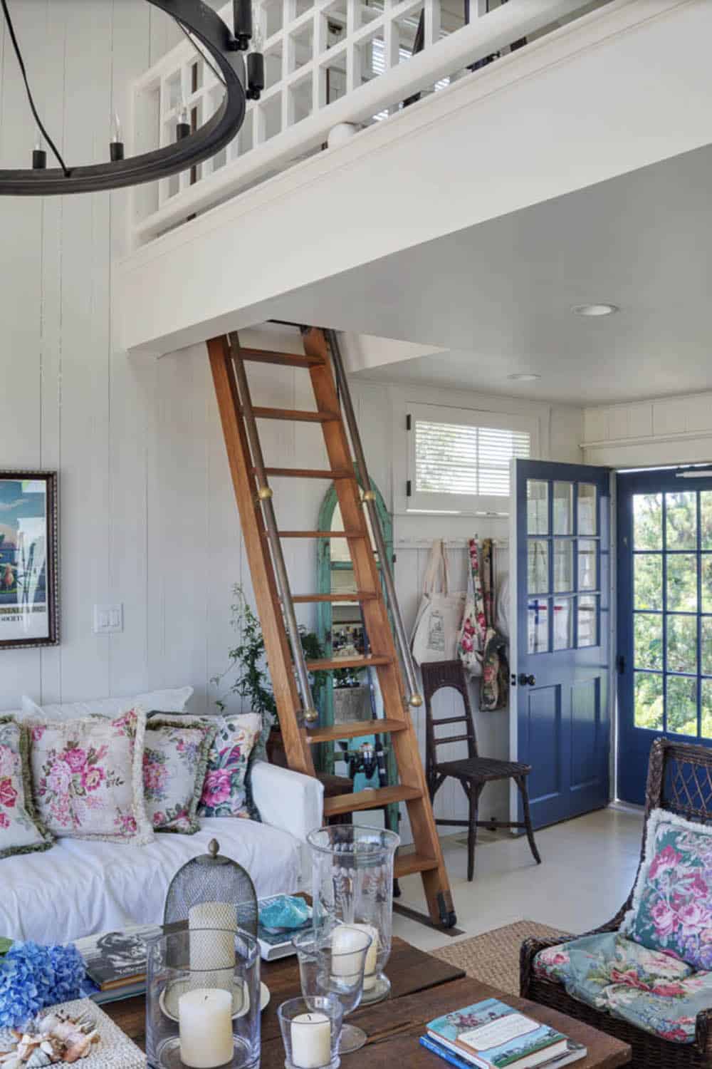Living room with rolling library ladder to loft, white shiplap walls, floral sofa, and blue front door