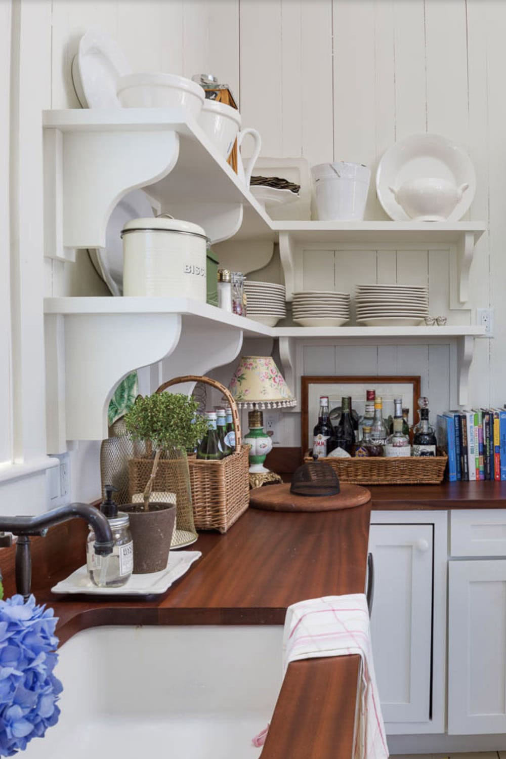 Cottage kitchen corner with open white shelving, wood countertops, wicker baskets, and bar setup