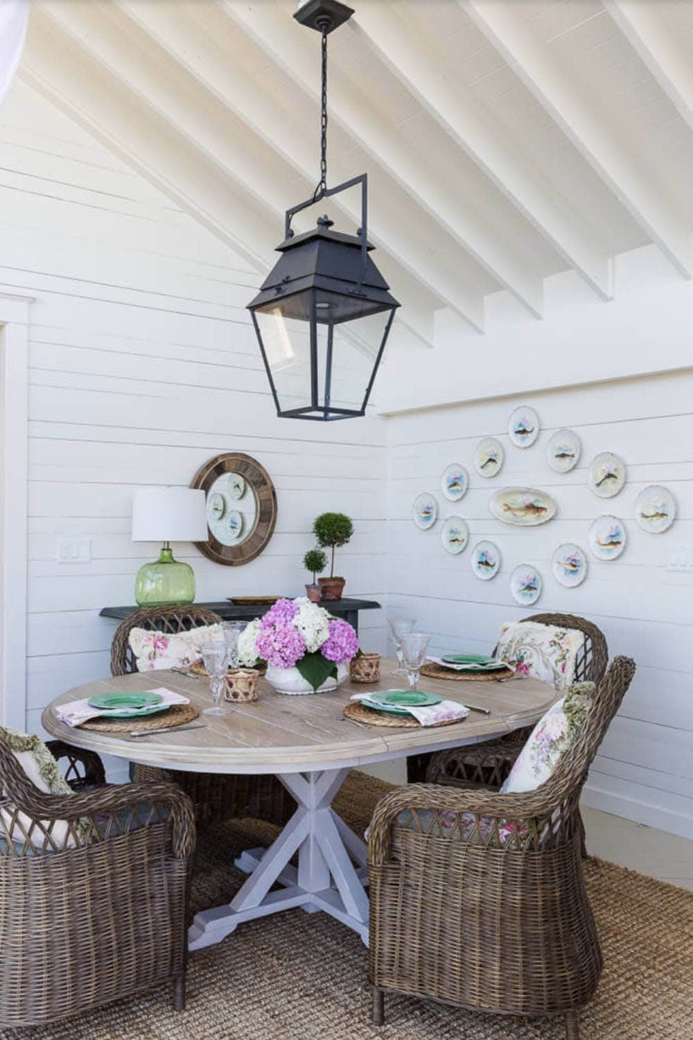 Screened porch dining area with round pedestal table, wicker chairs, lantern pendant, and fish plate wall display