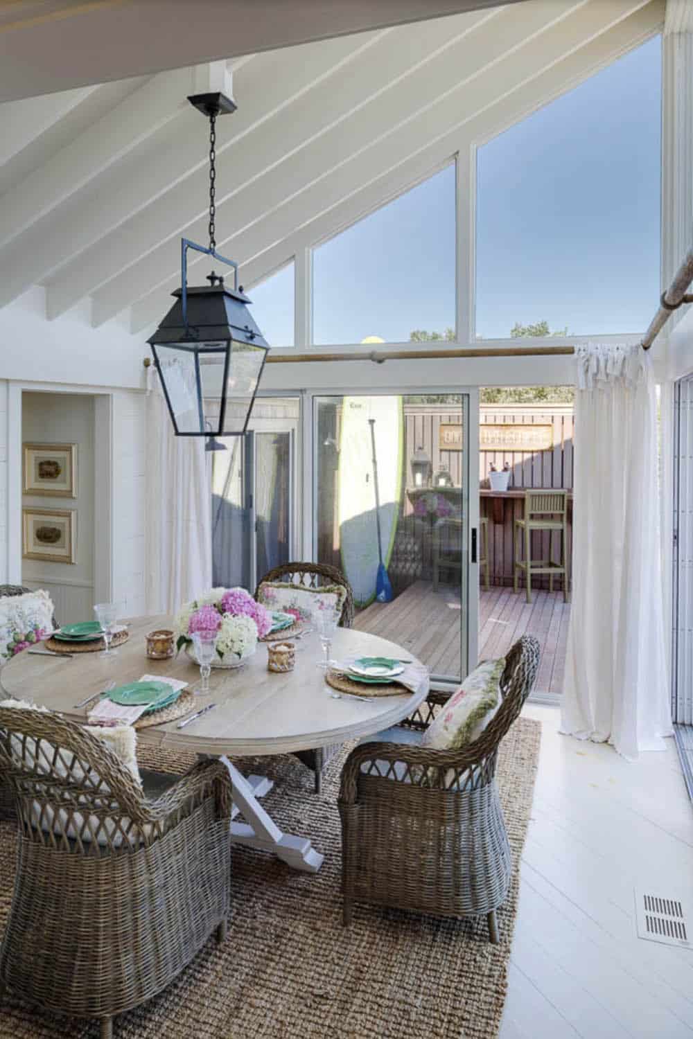 Sunroom dining area with vaulted ceiling, round wood table, wicker chairs, black lantern pendant, and sliding glass doors to deck