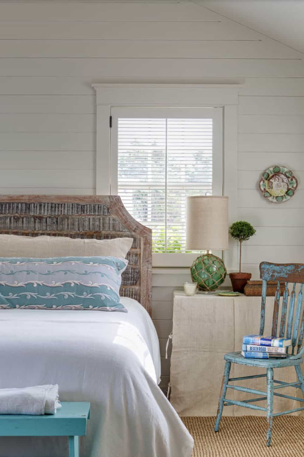 Coastal bedroom with carved wood headboard, embroidered blue pillow, rope lamp, and distressed blue chair