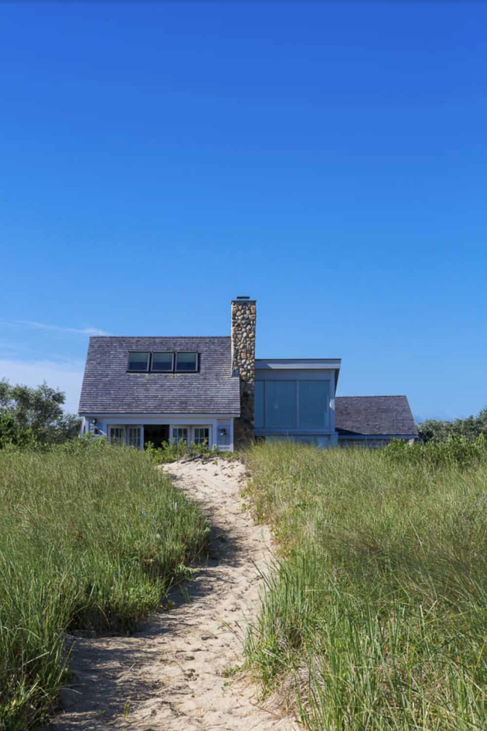 Sandy dune path leading to cedar shingle beach house with stone chimney and modern glass addition