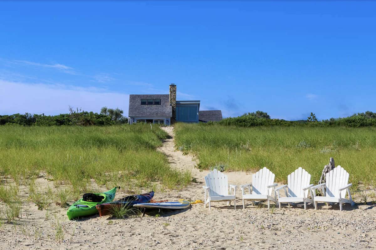 White Adirondack chairs and kayaks on sandy beach with cedar shingle cottage in background