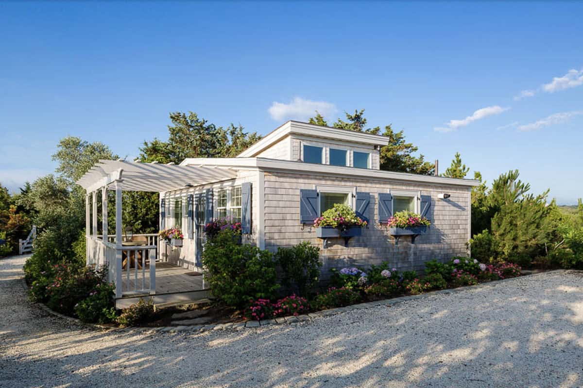 Cedar shingle guest cottage with white pergola, blue shutters, colorful window boxes, and gravel driveway