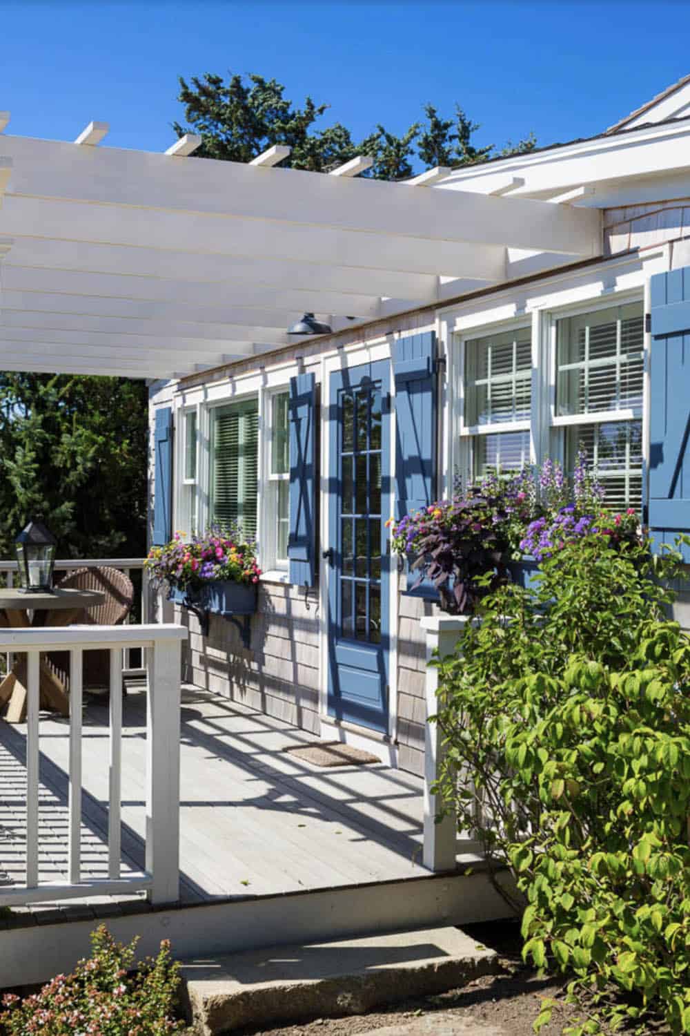 Cottage facade with white pergola, blue shutters, flower-filled window boxes, and white deck railing