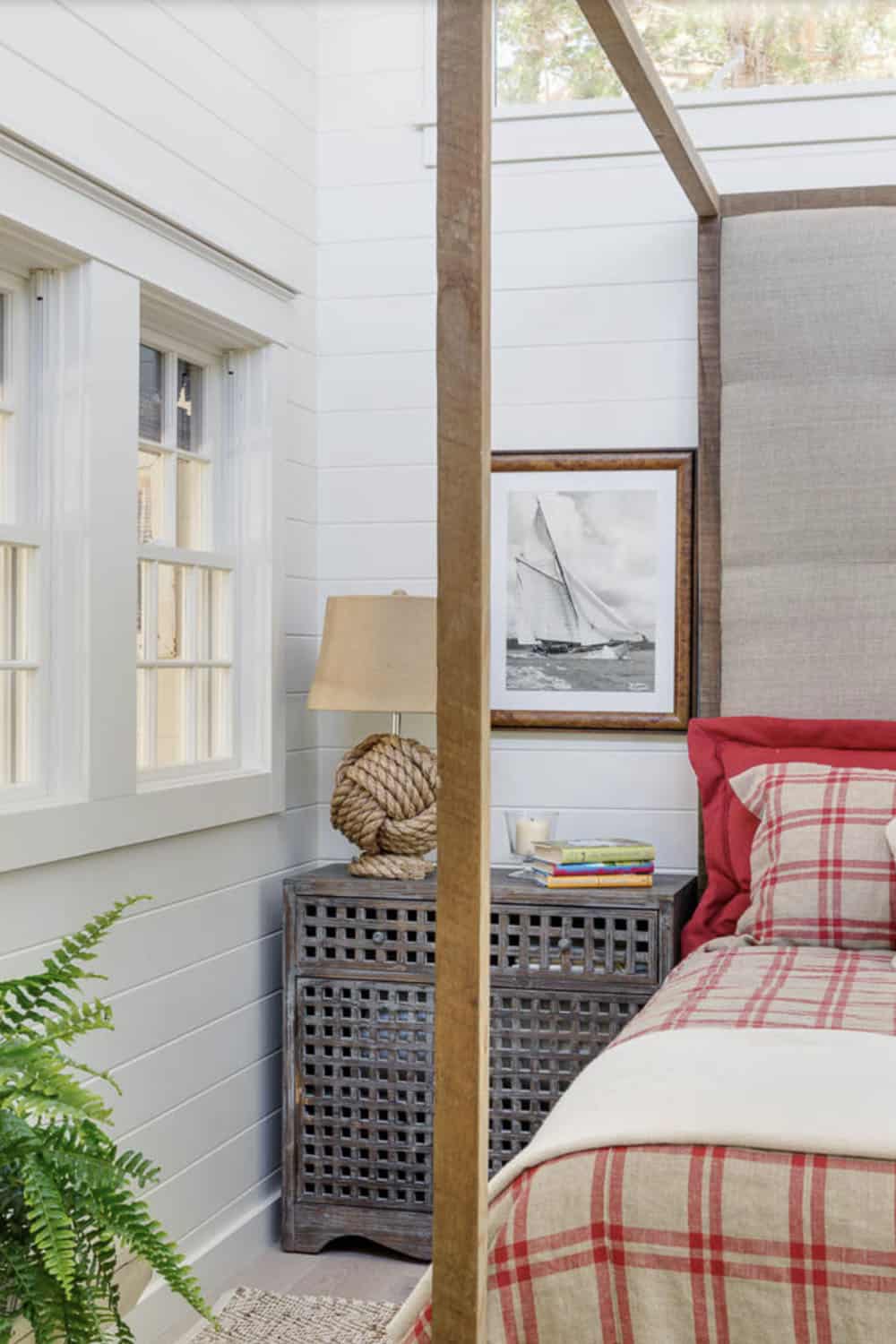 Cottage bedroom detail with rope-knot lamp, carved nightstand, sailboat print, and red plaid bedding