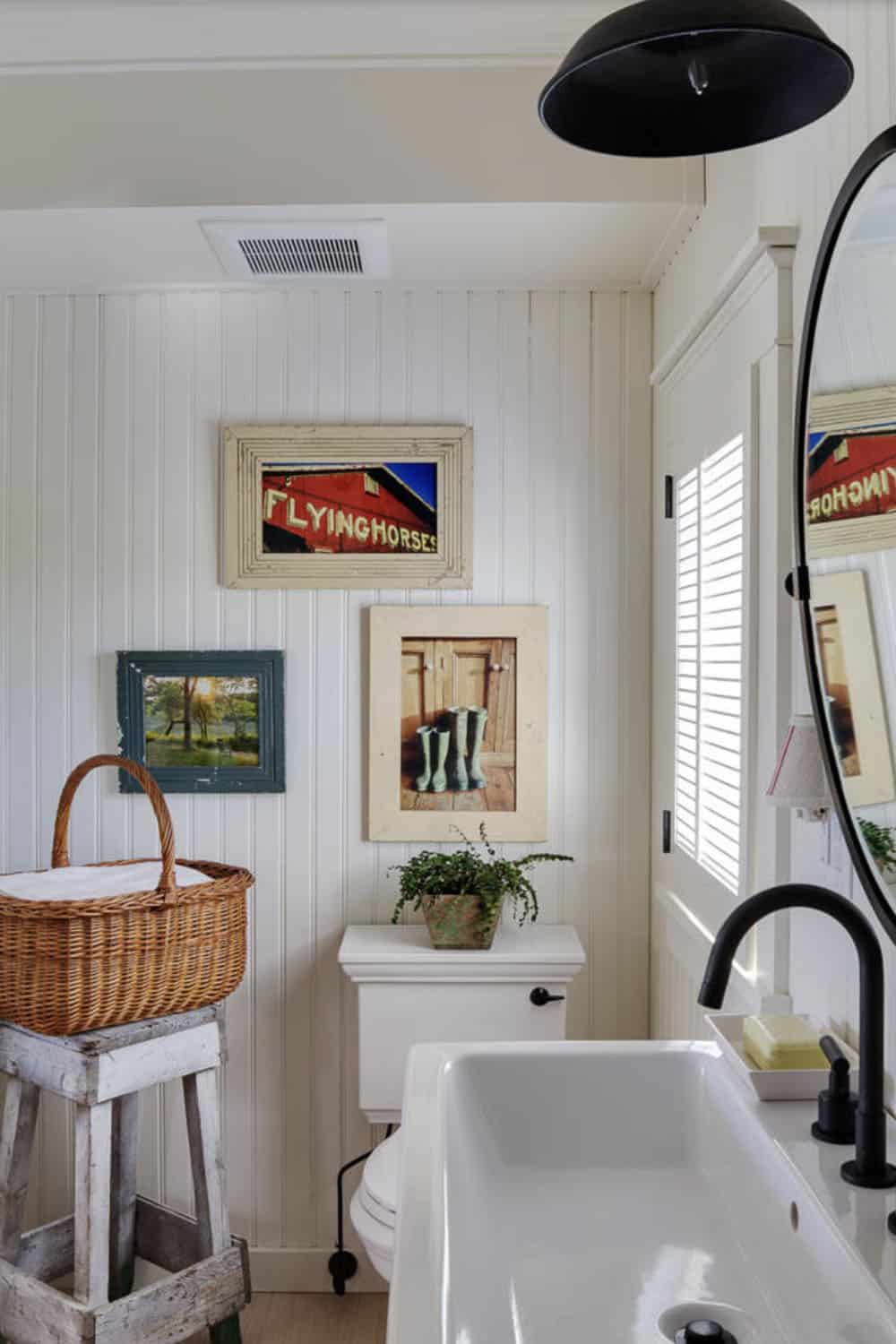Cottage bathroom with beadboard walls, black pendant light, round mirror, farmhouse sink, and whimsical local art