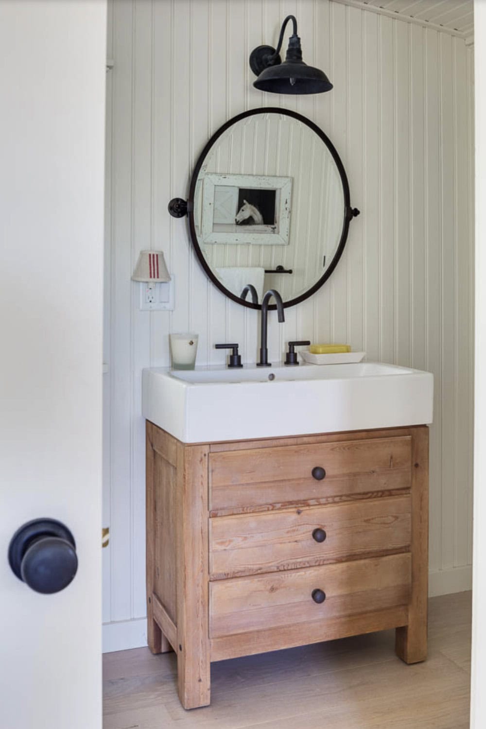 Cottage bathroom vanity with reclaimed wood dresser base, round black mirror, and barn sconce light